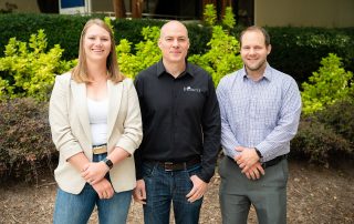 Three people standing in a row with greenery in the background.